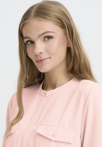 Blusa rosa chiaro con scollo rotondo, due tasche sul petto e dettagli con bottoni, realizzata in un tessuto liscio con una vestibilità leggermente ampia.