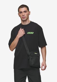 Borsa a tracolla nera realizzata in materiale resistente. Presenta una chiusura con zip, una tracolla regolabile e un logo verde brillante. Design compatto.