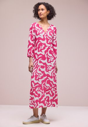 Kvinde med krøllet hår iført en lang kjole med pink og hvide mønstre og hvide sneakers, står mod en ensfarvet beige baggrund.