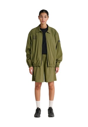 Vrouw staand, draagt een olijfgroene jas met ritssluiting en bijpassende knielange shorts, een zwart shirt, witte sokken en zwarte sneakers.