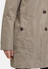 Beige vattentålig kappa med en framfickav, knapplister och en slät, mjuk textur. Sticksömmar längs kanterna.