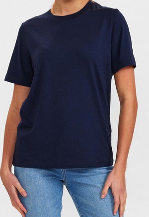 Vrouw draagt een effen marineblauw T-shirt met korte mouwen en lichtblauwe spijkerbroek, handen rustend nabij de heupen tegen een effen achtergrond.