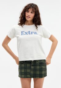 Bela bombažna majica s kratkimi rokavi in modrim napisom "Extra." na sprednji strani, v kombinaciji z zelenim karo denim krilom. Ohlapen kroj.