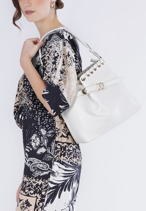 Femme en robe à motifs portant un grand sac à main en cuir blanc avec détail boucle sur l'épaule, sur un fond clair.