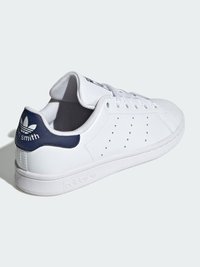 White leather sneaker with navy accents, perforated sides, round toe, and flat sole. Features branded heel and tongue. Minimalist design.