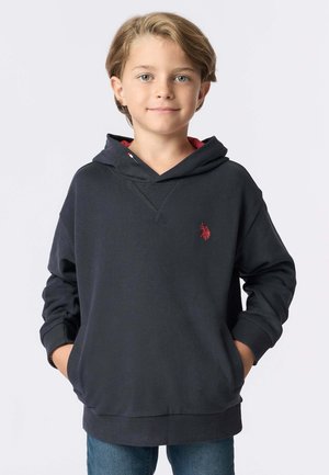 Ragazzo sorridente con capelli castano chiaro, indossa un hoodie blu scuro con un logo rosso e jeans blu, con le mani nelle tasche, in piedi su uno sfondo bianco.