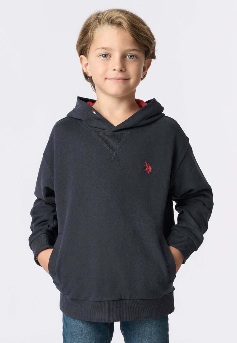 Ragazzo sorridente con capelli castano chiaro, indossa un hoodie blu scuro con un logo rosso e jeans blu, con le mani nelle tasche, in piedi su uno sfondo bianco.