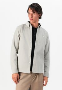 Young man wearing a light gray button-up shirt over a black polo and beige pants, looking sideways against a plain white background.