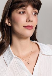 Złoty naszyjnik z zawieszką w kształcie półksiężyca. Modelka ma na sobie białą, teksturowaną koszulę. Miękkie światło podkreśla detale oraz blask naszyjnika.