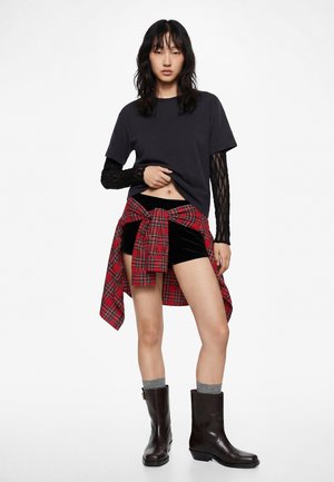 Kvinde iført en sort kortærmet skjorte over en sort blonder langærmet top, sorte shorts, en rødternet skjorte bundet om livet, grå sokker og mørke støvler.