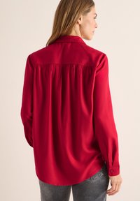 Rød silke bluse med en løs pasform, knaplukning ved ærmerne, og samlet ryg. Har en klassisk krave og en glat, skinnende tekstur.