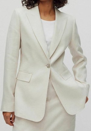 Mujer con un blazer entallado de color crema con un botón abrochado sobre una blusa blanca, combinado con una falda a juego.