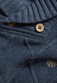 Donkerblauw gebreid sweater met een textuurpatroon, voorzien van een geribbelde kraag en twee knopen met houtlook. Inclusief een geborduurd accent.