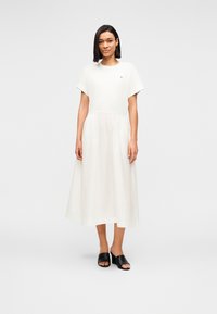 Γυναίκα που στέκεται φορώντας λευκό μεσομήκης φόρεμα με κοντά μανίκια και μαύρα mule με ανοιχτά δάχτυλα, μπροστά σε απλό ανοιχτό γκρι φόντο.