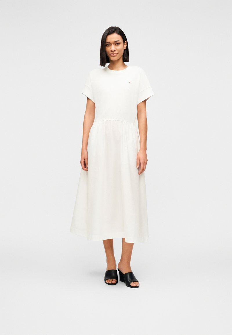 Γυναίκα που στέκεται φορώντας λευκό μεσομήκης φόρεμα με κοντά μανίκια και μαύρα mule με ανοιχτά δάχτυλα, μπροστά σε απλό ανοιχτό γκρι φόντο.