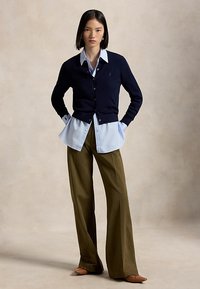 Frau trägt einen marineblauen Cardigan über einem hellblauen Hemd mit olivgrünen, weit geschnittenen Hosen und braunen Ballerinas, steht mit den Händen in den Taschen.