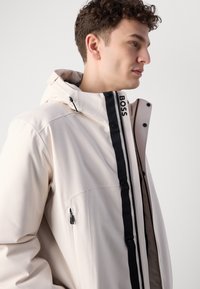 Lyse beige jakke med lynlås foran, sorte detaljer og hætte. Tekstureret stof har sidelommer og et logo på kraven.
