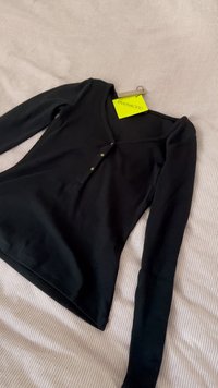 Maglia nera a maniche lunghe con scollatura a barca, caratterizzata da tre dettagli bottoncini, realizzata in tessuto morbido, distesa su una superficie a righe chiare.
