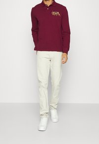 Burgundy långärmad polo med en broderad design, i kombination med ljusa byxor och vita sneakers.