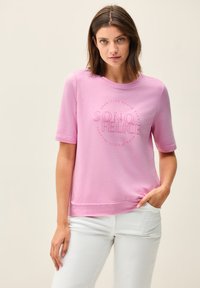 Chemise en mélange de coton rose à manches courtes, ornée d'un graphique circulaire avec l'inscription "SONO FELICE" au centre, associée à un pantalon blanc.