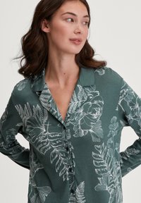 Camicia verde a maniche lunghe con colletto, caratterizzata da un motivo floreale e foglioso bianco, con una texture morbida.