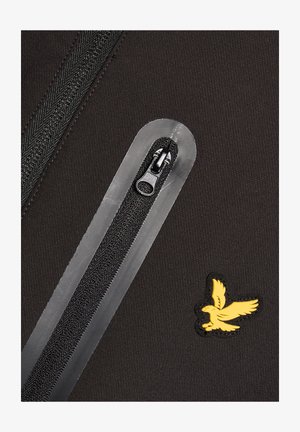 Zwart stof met een verticale waterdichte rits en een geel adelaarlogo geborduurd nabij de rechteronderhoek.