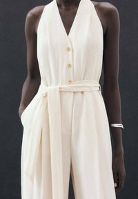 Filière sans manches crème avec un décolleté en V profond, une fermeture à boutons sur le devant, une ceinture à la taille et des poches latérales. Texture de tissu lisse et léger.