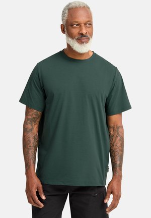 T-shirt verde scuro realizzata in tessuto morbido. Maniche corte, scollo rotondo e vestibilità comoda. Mostra tatuaggi visibili sulle braccia del modello.