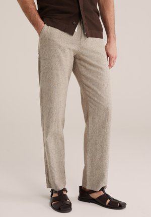 Man draagt beige getextureerde rechte broek, donkerbruin overhemd met knopen en donkerbruine lederen sandalen met gespen.