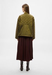 Sötét olívazöld, steppelt bomber dzseki sima felülettel, lekerekített gallérral és hátul lévő yoke-kal, barna pliszírozott szoknyával párosítva.