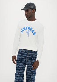 White long-sleeve shirt with blue "ICECREAM" graphic. Dark denim pants featuring repeated "ICECREAM" text pattern, black cap with leather texture.