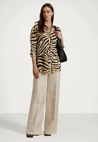 Γυναίκα με πουκάμισο με σχέδιο ζέβρας, φαρδιά κρεμ παντελόνια, μαύρη τσάντα ώμου, που στέκεται μπροστά σε απλό φόντο.