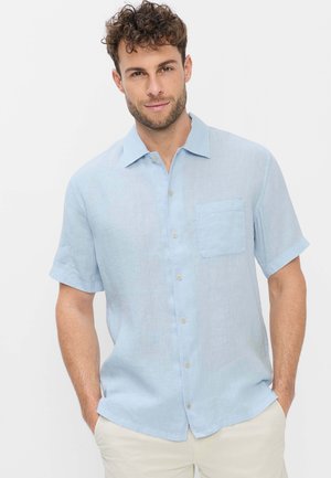 Ung mand med krøllet hår iført en lyseblå kortærmet skjorte og beige bukser, hænderne i lommerne, mod en ensfarvet baggrund.