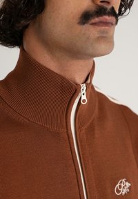 Mand med overskæg iført en brun lynlås-sweater med ribbet krave og hvidt broderet logo på brystet.