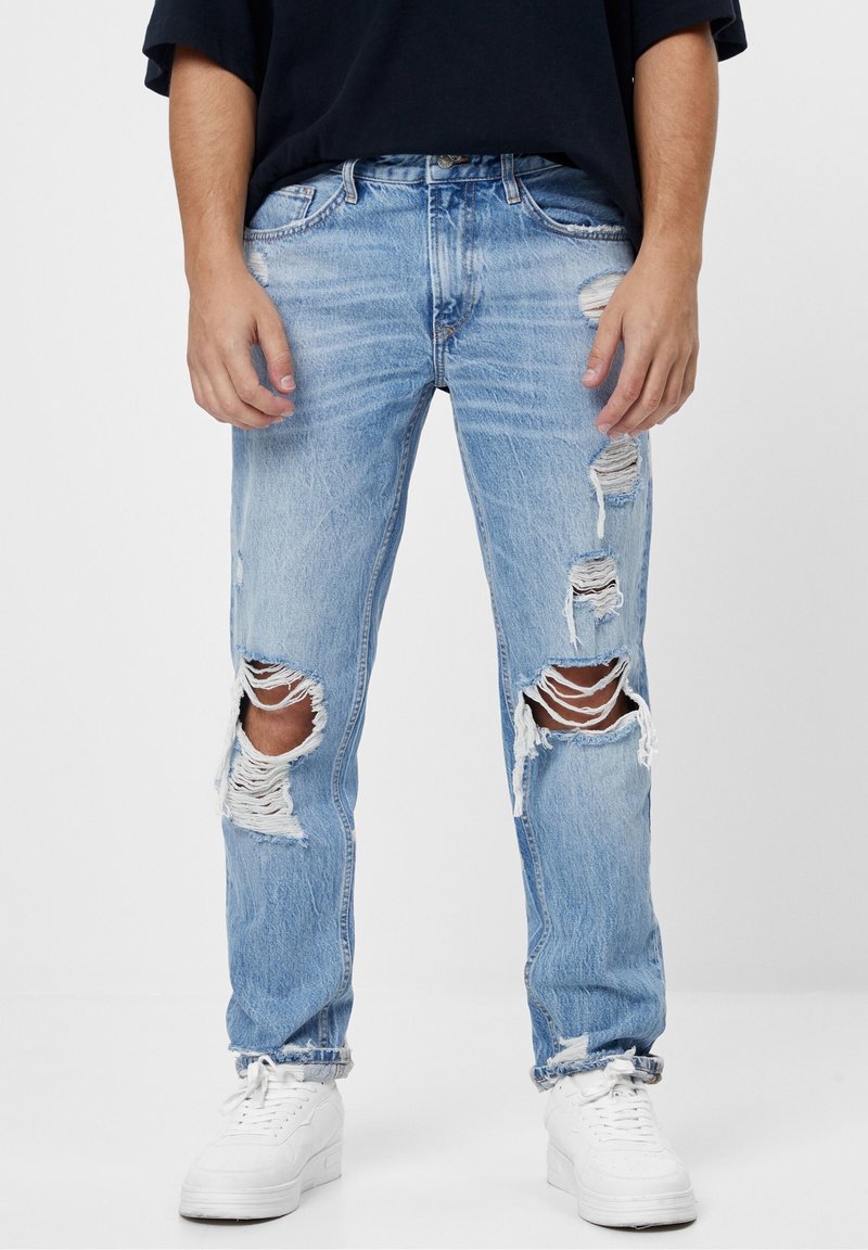 Ljusblå slitna jeans med revor vid knän och lår, fransiga kanter, vid passform, lätt blekning, kombinerade med vita sneakers, bomullsmaterial.