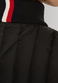 Giacca a vento nera con colletto a costine e strisce rosse, bianche e blu navy. Superficie texture con motivo trapuntato e cuciture visibili.