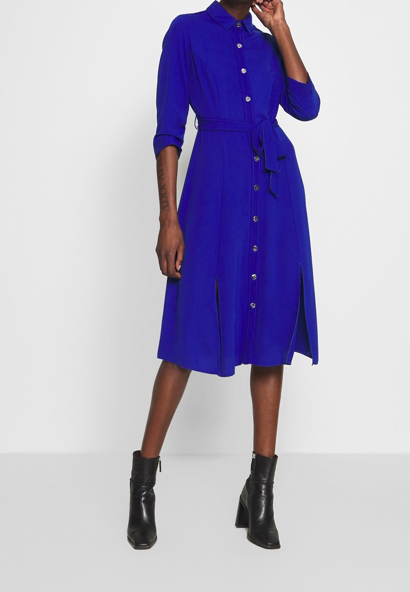 Blå skjortklänning med långa ärmar, knappar framtill och knytning i midjan. Har sidospjäll och en slät textur. Bärs med svarta ankelstövlar.