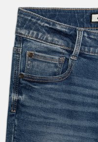 Los pantalones de mezclilla presentan un lavado azul medio con un desgaste sutil, costuras de contraste, un bolsillo delantero y una etiqueta de logo en la cinturilla.