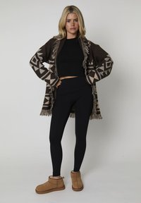 Blonde vrouw staat met handen op de heupen, gekleed in een zwarte crop top, zwarte leggings, een bruine geprinte franjes cardi en tan enkellaarsjes.