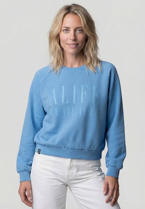 Blonde Frau, die einen hellblauen Pullover mit dem Schriftzug "ALIFE" und weiße Hosen trägt, vor einem einfarbigen, hellgrauen Hintergrund steht.