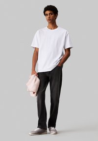 Camiseta de algodón blanca con mangas cortas y un logotipo sutil, combinada con jeans oscuros, sosteniendo un suéter de punto rosa claro y llevando zapatillas grises.