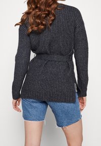Mörkgrå stickad cardigan med bälte i midjan, långa ärmar och sidoflikar. Bärs över blå denimshorts med fransad nederkant.