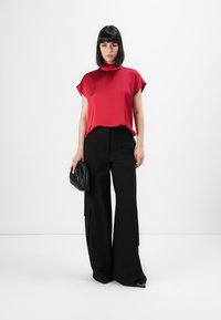 Femme portant un chemisier en satin rouge et un pantalon noir à jambes larges, tenant une pochette noire, se tenant devant un fond blanc uni.
