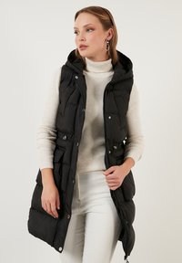 Čierny puffer vest s kapucňou, so zipsom vpredu a cvokmi. Má bočné vrecká a prešívanú štruktúru. Nosený cez biely rebrovaný sveter.