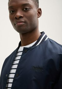 Chaqueta bomber de color navy con acentos de franjas blancas en el cuello, que cuenta con un cierre a presión y un logotipo bordado "DENIM TOM TAILOR".