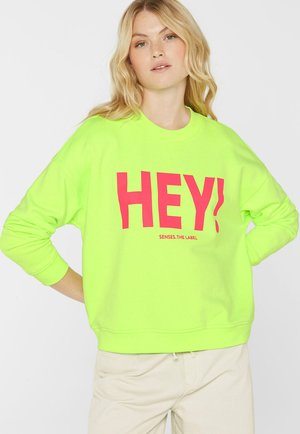 Blonde Frau trägt ein leuchtend gelb-grünes Sweatshirt mit großem rotem "HEY!"-Schriftzug, steht mit einer Hand in der Hüfte.