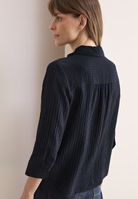 Blusa nera in tessuto texturizzato con colletto, pieghe posteriori e polsini con bottoni. Il design presenta righe verticali e una vestibilità morbida.