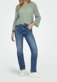 Pull vert clair en tricot avec un col rond et des manches cloche, associé à un jean bleu droit et des baskets blanches.