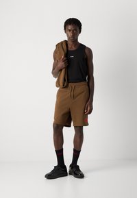 Tânăr îmbrăcat cu un tricou fără mâneci negru, pantaloni scurți maro, ținând un hanorac maro pe umăr, cu șosete negre și adidași negri, pe un fundal alb.