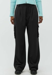 Schwarzfarbene Cargohosen mit weitem Bein aus Stoff, mit seitlichen Taschen und Falten. Kombiniert mit einer hellblauen gerippten Strickjacke.